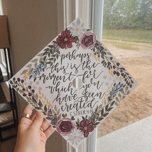 May include: A graduation cap with floral accents and the quote "perhaps this is the moment for which you have been created" from Esther 4:14. The cap features burgundy, white, and blue flowers and green foliage. The cap is held up near a window.