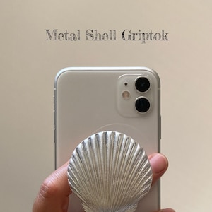 May include: A silver metal shell phone grip attached to a silver smartphone. The grip is in the shape of a scallop shell. The text "Metal Shell Griptok" is above the phone.