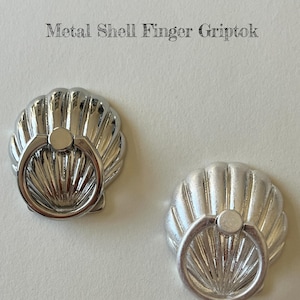 May include: Two silver metal shell-shaped finger grip phone holders. Each has a ring for holding the phone. The text "Metal Shell Finger Griptok" is at the top of the image.