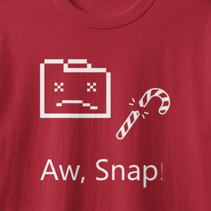May include: A red t-shirt featuring a white graphic of a pixelated sad computer folder icon next to a broken candy cane. Below the graphic, the text reads Aw, Snap! in white lettering. The design is centered on the front of the garment.