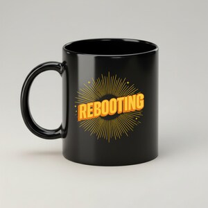 May include: Black ceramic mug with a curved handle. The mug features the word "REBOOTING" in orange, outlined in yellow, set against a yellow burst design. The mug is designed for hot beverages.