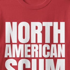 May include: A red t-shirt featuring the text NORTH AMERICAN SCUM printed in large, bold, white capital letters across the chest. The shirt has a standard crew neckline and a smooth fabric texture, with the graphic centered on the front.
