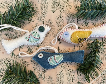 Puede incluir: Tres adornos de pájaros de fieltro con diferentes patrones y colores. Los pájaros son blancos, amarillos y azules con una cuerda blanca para colgar.