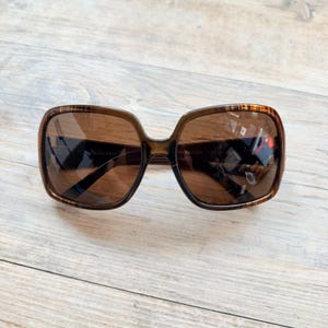 May include: Brown square sunglasses with a tortoiseshell pattern on the upper frame. The dark lenses reflect light. The sunglasses are on a light wooden surface. The text "Thirty-First" is visible on the left lens.