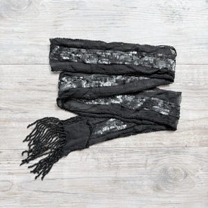 May include: A long black scarf made of sheer fabric featuring horizontal bands of shimmering silver sequins. The edges of the scarf are finished with delicate black beadwork, and one end terminates in a dense fringe of long black beaded tassels.