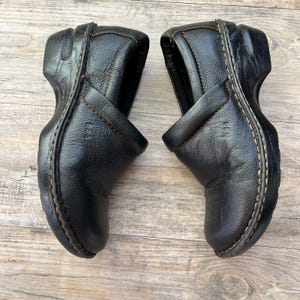 May include: Pair of black leather clogs with a rounded toe and a slip-on design. The shoes have a textured leather upper, visible stitching, and a thick, dark sole. The clogs are displayed on a light brown wooden surface.