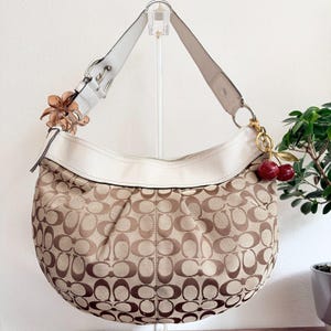 May include: A hobo-style shoulder bag featuring a beige and brown monogram pattern on the body with a white leather trim and strap. The bag includes a brown flower-shaped charm on one side and a gold-toned cherry charm on the other side.