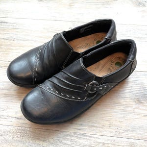 May include: Pair of black leather slip-on shoes with white stitching details. The shoes have a rounded toe and a small buckle accent. The insoles have a leaf design. The shoes are on a light wood surface.