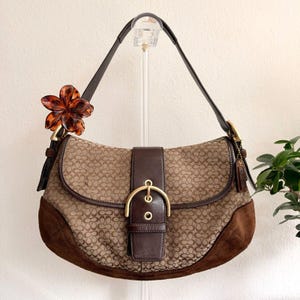 May include: A brown shoulder bag featuring a repeating logo pattern on the main body, dark brown leather trim, and suede side panels. The bag includes a central buckle closure with gold-tone hardware and a decorative tortoiseshell flower charm.