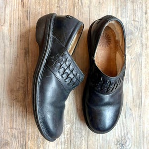 May include: A pair of black leather clogs with a woven design on the vamp. The clogs have a rounded toe and a low heel. The interior of the clogs is a light brown colour. The clogs are on a wooden surface.