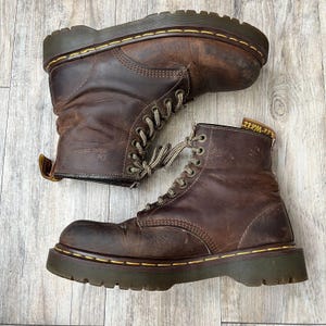 Puede incluir: Un par de botas Dr. Martens de cuero marrón con diseño de cordones. Las botas tienen una suela gruesa de color verde oscuro con costuras amarillas y un cuello acolchado. El logotipo de Dr. Martens es visible en el lateral de la bota.