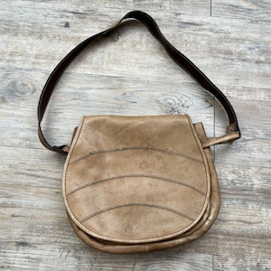 May include: Tan leather shoulder bag with a brown strap. The bag has a curved shape with horizontal stitched details across the front. The bag is sitting on a wooden surface.