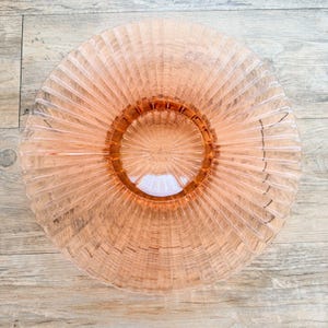 Op de afbeelding: Een ronde, doorschijnende roze glazen schaal met een opvallend zonnestraalpatroon met stralende ribbels die zich uitstrekken van het midden naar de geschulpte buitenrand. De schaal heeft een gladde, ronde voet en een brede opening.