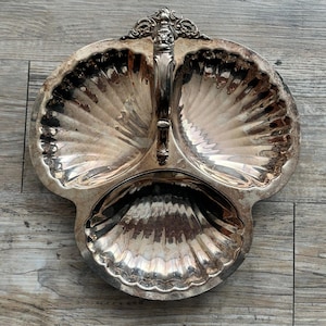 May include: A silver-colored, three-section serving dish with a shell-like design. The dish has a handle with ornate detailing and is likely made of metal. The sections are divided into three distinct areas, perfect for serving appetizers or condiments.