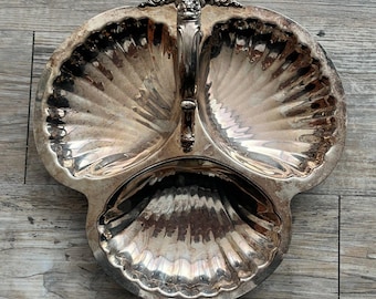Pode incluir: Uma travessa de servir prateada com três secções e um design em forma de concha. A travessa tem uma pega com detalhes ornamentados e é provavelmente feita de metal. As secções são divididas em três áreas distintas, perfeitas para servir aperitivos ou condimentos.