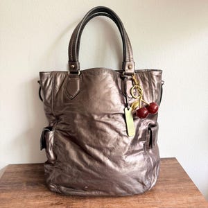 May include: A metallic bronze tote bag with two top handles and two side pockets. A gold-toned metal tag and a dangling charm featuring two red cherries with a gold stem are attached to the right handle base, adding detail to the textured surface.