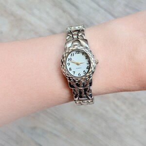 Peut inclure: Une montre à quartz argentée dotée d'un cadran blanc avec des chiffres arabes noirs et des aiguilles dorées. Le boîtier et le bracelet présentent une finition texturée et alvéolée, avec de petites pierres claires incrustées dans les maillons.