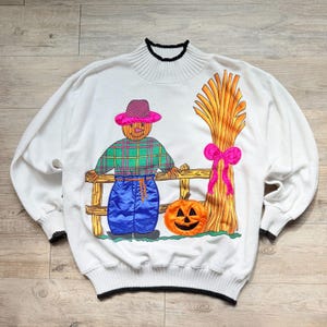 May include: A white long-sleeve sweatshirt featuring a mock neck with black trim. The front displays a colorful applique of a scarecrow in a plaid shirt and blue pants, standing by a wooden fence with a pumpkin and a tall sheaf of wheat.