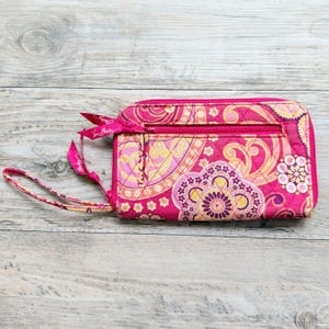 May include: A rectangular quilted wallet in shades of hot pink, yellow, and orange with a paisley and floral pattern. It features a zippered exterior pocket, a matching fabric wrist strap, and a full-length zipper closure along the top edge.