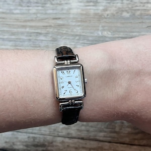 Puede incluir: Un reloj rectangular plateado con esfera blanca y números negros. El reloj tiene una correa de cuero negro. La esfera del reloj dice "CARRAGE QUARTZ".