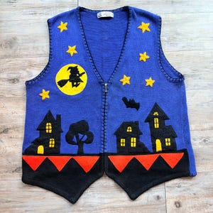 Puede incluir: Un chaleco de punto azul con cierre de cremallera frontal y un diseño con temática de Halloween. La prenda muestra estrellas amarillas, una luna amarilla con la silueta de una bruja negra, casas negras con ventanas amarillas, un árbol negro, un murciélago negro y triángulos naranjas.