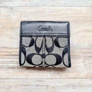 May include: A black and gray bi-fold wallet featuring the signature Coach logo pattern on a textured fabric body. The wallet has a solid black leather top panel with the word Coach embossed in a script font across the center front.