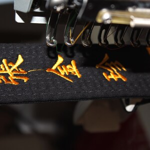 May include: Close-up of a black belt with gold embroidery that reads "黑 - 跆拳 道". The belt is being embroidered by a machine.