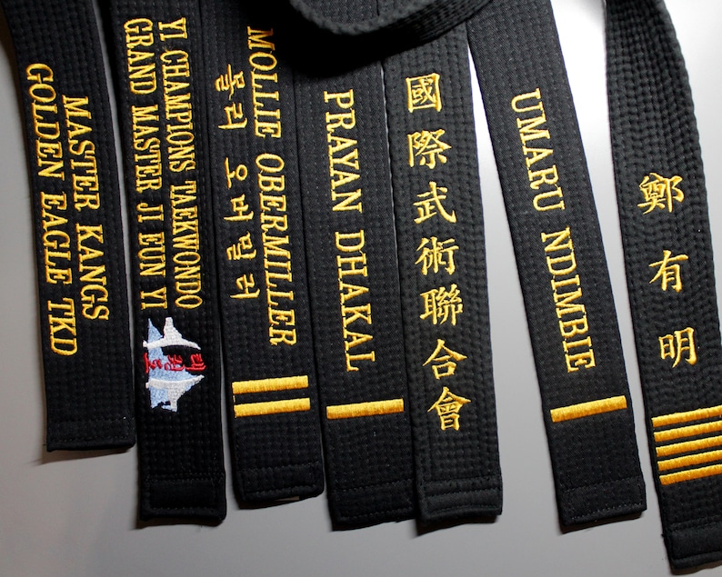 May include: A close-up of several black martial arts belts with gold embroidered names and text. The names include "Master Kangs", "Golden Eagle TKD", "Yi Champions Taekwondo", "Grand Master Ji Eun Yi", "Mollie Obermiller", "Prayan Dhakal", "Umar Ndmbie", "International Martial Arts Federation", and "Zheng Youming".