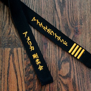 May include: Black martial arts belt with gold embroidered Japanese characters. The characters on the belt read "Aikido" and "Shukokai".