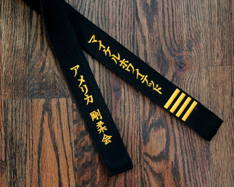 May include: Black martial arts belt with gold embroidered Japanese characters. The characters on the belt read "Aikido" and "Shukokai".