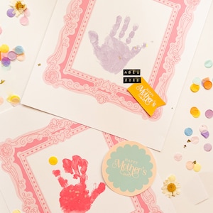 Puede incluir: Dos tarjetas hechas a mano para el D&iacute;a de la Madre con huellas de manos dentro de marcos decorativos rosas. Una huella es morada, la otra roja. Las tarjetas tienen los nombres "Abel" y "Ellis" con el a&ntilde;o "2023". Confeti y una pancarta de "Happy Mother's Day" est&aacute;n esparcidos.