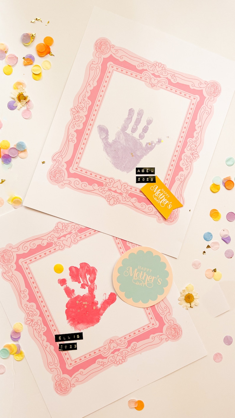 Puede incluir: Dos tarjetas hechas a mano para el D&iacute;a de la Madre con huellas de manos dentro de marcos decorativos rosas. Una huella es morada, la otra roja. Las tarjetas tienen los nombres "Abel" y "Ellis" con el a&ntilde;o "2023". Confeti y una pancarta de "Happy Mother's Day" est&aacute;n esparcidos.