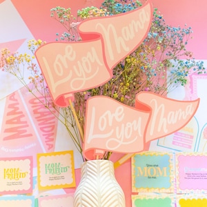 Puede incluir: Una colecci&oacute;n de tarjetas y decoraciones para el D&iacute;a de la Madre en tonos rosa y blanco. La pieza central es un jarr&oacute;n con flores coloridas y pancartas que dicen "Love you Mama". Otras tarjetas dicen "Mom-Friend" y "You are MOM to me."