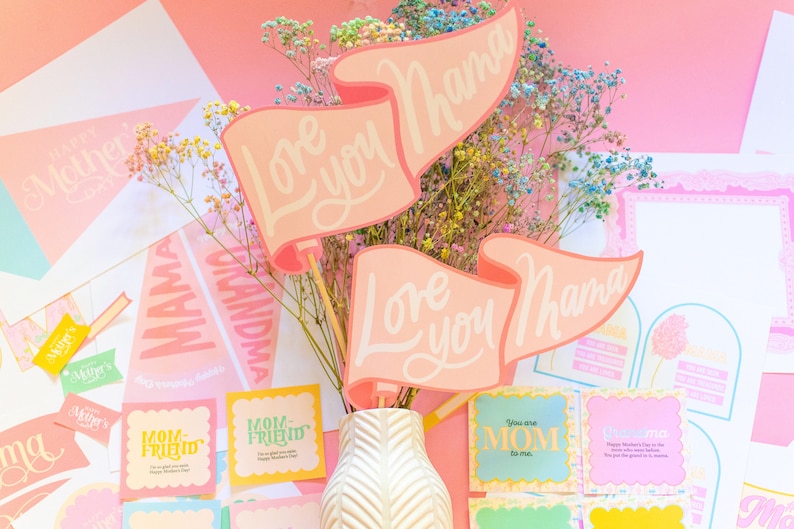 Puede incluir: Una colecci&oacute;n de tarjetas y decoraciones para el D&iacute;a de la Madre en tonos rosa y blanco. La pieza central es un jarr&oacute;n con flores coloridas y pancartas que dicen "Love you Mama". Otras tarjetas dicen "Mom-Friend" y "You are MOM to me."