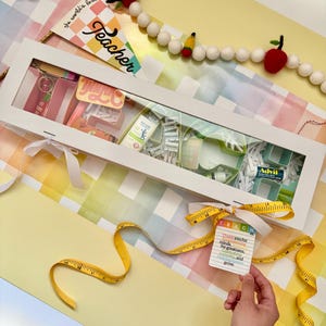 May include: A white rectangular gift box filled with various items, including a keychain, notepad, and Advil. A yellow measuring tape and a tag that says "TEACH" are also included. The box is tied with a white ribbon and sits on a colorful background.