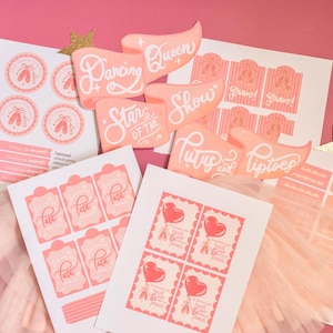 May include: A collection of pink and white ballet-themed party supplies, including stickers, labels, and decorative cutouts with phrases like "Dancing Queen" and "Star of the Show." Gold star-shaped confetti adds a festive touch.