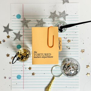 May include: A yellow notepad with the text "the TORTURED teachers department" on the front. The notepad is on a sheet of lined paper with silver glitter stars and blue and gold confetti. There is a magnifying glass and a disco ball on the paper.