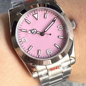 May include: A silver wristwatch with a pink face and white markings. The watch has a silver metal band with the number "904L" printed on it.
