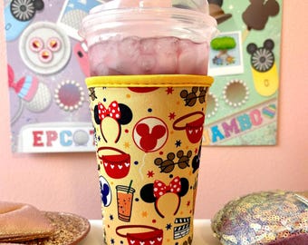 May include: A yellow and red neoprene cup holder with a Mickey Mouse theme. The cup holder features images of Mickey Mouse ears, Mickey Mouse balloons, and Mickey Mouse hats. The cup holder is designed to fit a standard-sized cup.