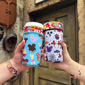 May include: Two colorful reusable cup sleeves with a Mickey Mouse theme. One sleeve features a blue background with Mickey Mouse, Minnie Mouse, and various Disney treats. The other sleeve features a white background with Mickey Mouse, Minnie Mouse, and Halloween-themed icons.