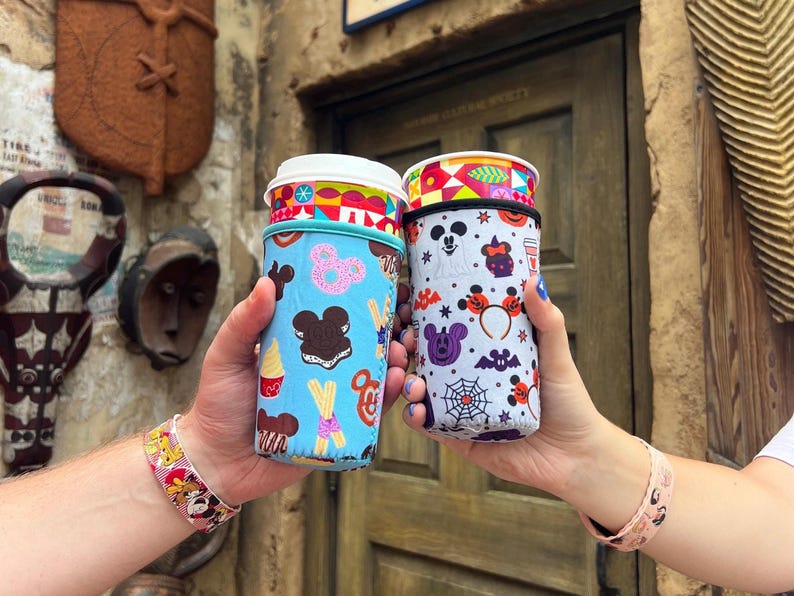May include: Two colorful reusable cup sleeves with a Mickey Mouse theme. One sleeve features a blue background with Mickey Mouse, Minnie Mouse, and various Disney treats. The other sleeve features a white background with Mickey Mouse, Minnie Mouse, and Halloween-themed icons.