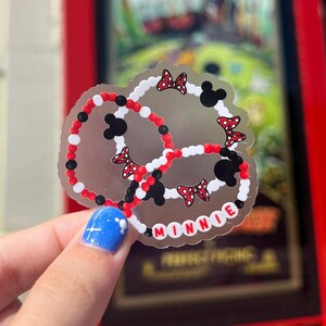 May include: A silver sticker with a Minnie Mouse design. The sticker features three red, white, and black bracelets with Minnie Mouse ears and bows. The word "MINNIE" is spelled out in white beads around the edge of the sticker.