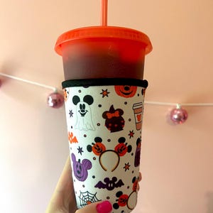 May include: A white reusable cup sleeve with a Halloween-themed pattern featuring Mickey Mouse, Minnie Mouse, and other Disney characters. The sleeve has a black band at the top and bottom. The cup sleeve is on a red cup with a straw.