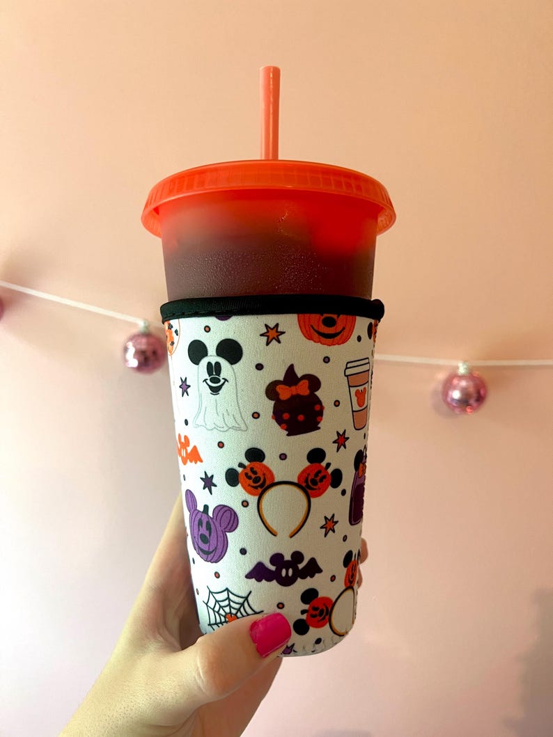 May include: A white reusable cup sleeve with a Halloween-themed pattern featuring Mickey Mouse, Minnie Mouse, and other Disney characters. The sleeve has a black band at the top and bottom. The cup sleeve is on a red cup with a straw.