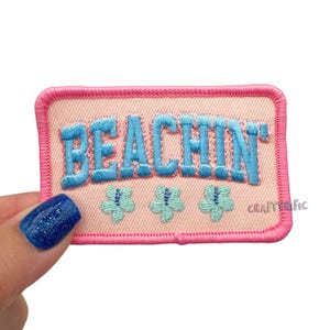 May include: A rectangular patch with the word "BEACHIN'" embroidered in blue, above three small blue and green flower designs. The patch has a pink border and a light peach background. The word "CRAFTERIFIC" is printed in the bottom right corner.