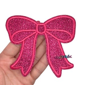 May include: A pink glitter bow applique with a stitched outline. The bow has a classic bow shape with two loops and two streamers. The text "CRAFTYRIFIC" is visible on the right side of the bow.