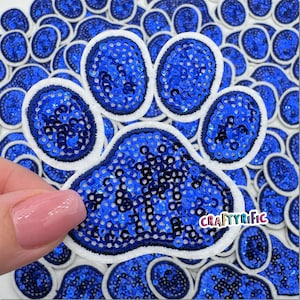 May include: Blue sequin paw print iron-on patch with white felt edging. The patch is surrounded by other paw print patches.