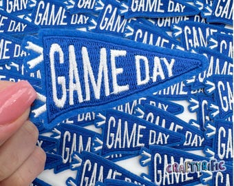 May include: A close-up of a pile of blue felt pennant patches with white embroidered text that reads "GAME DAY".