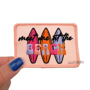 May include: A rectangular patch with a light pink background features three colorful surfboards. The text "meet me at the BEACH" is embroidered in black and colorful letters. The patch has a stitched border and the word "CRAFTYrific" is in the bottom right corner.