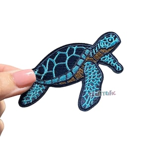 May include: Embroidered sea turtle patch in shades of blue and gold. The patch features a detailed design of a turtle with a textured shell and flippers. The patch is held by a hand against a white background.